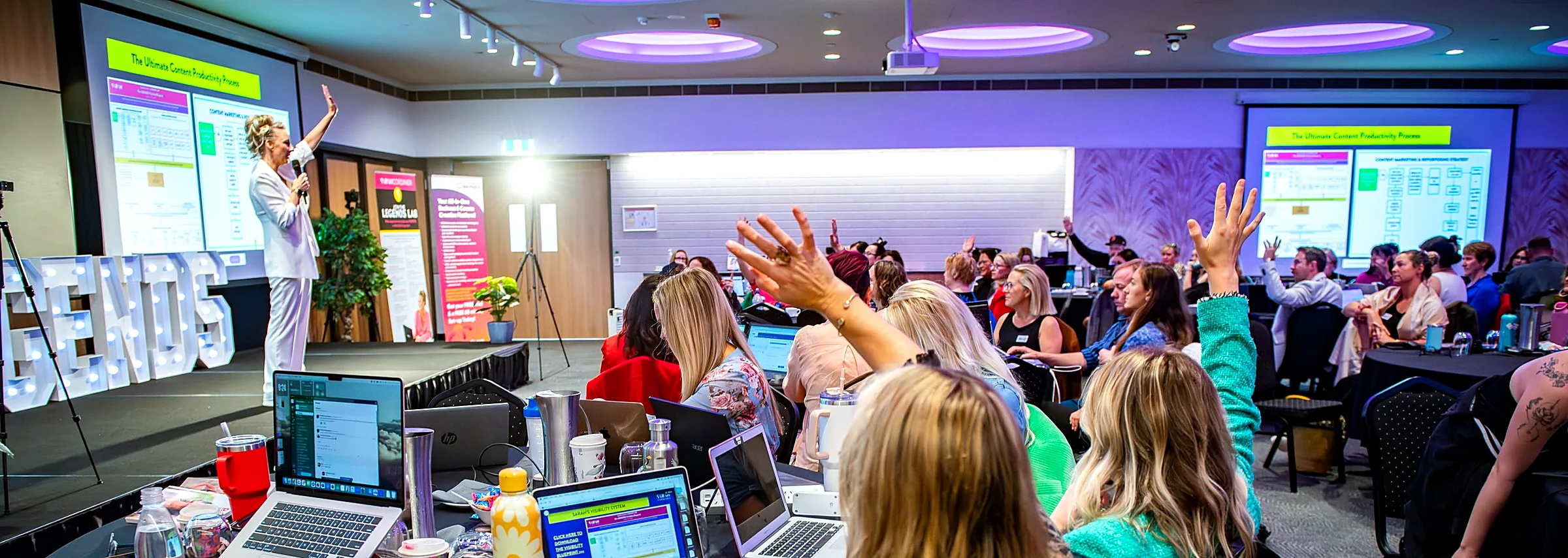 A full room of Legends Lab members with laptops open and hands raised while Sarah Cordiner teaches on stage
